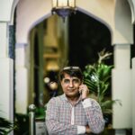 A South Asian man in casual attire talks on his mobile phone under a lit archway.